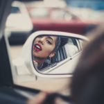 woman putting liquid lipstick on her lips while looking at vehicle's mirror during daytime