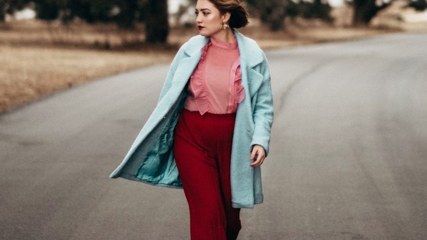 woman wearing teal coat and red pants walking on gray top road at day time