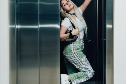 woman in green and white plaid dress shirt and black pants leaning on gray wall